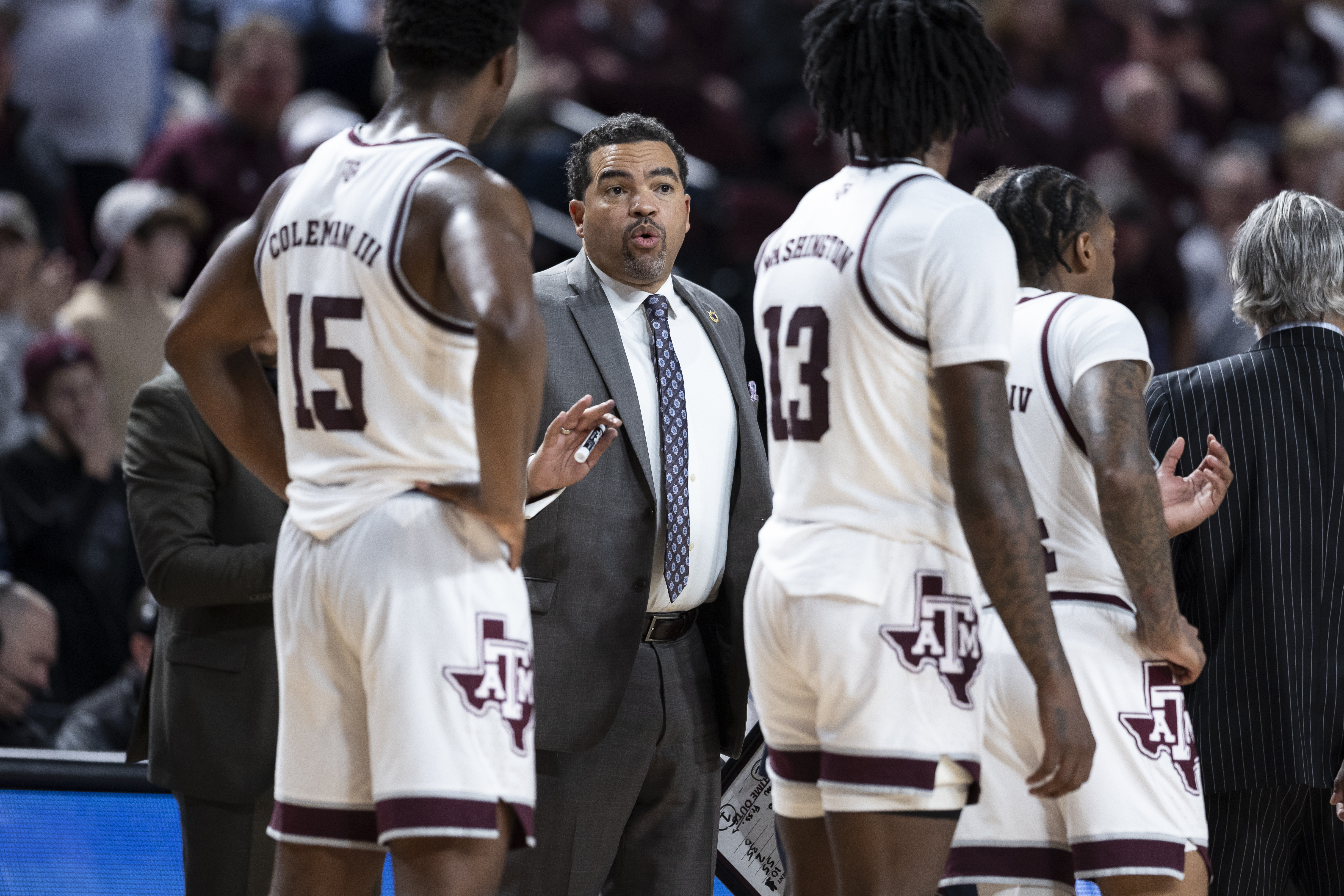 Vince Walden ’01, dressed in a gray suit and tie, speaks to Texas A&M men's basketball players during a game. He holds a marker and appears to be giving instructions during a timeout. Players in white Texas A&M uniforms with maroon trim surround him, and the arena crowd is visible in the background.